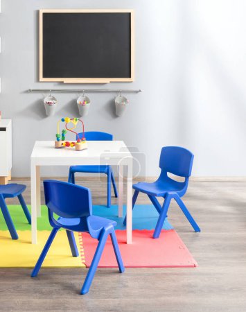 Helles und farbenfrohes Kindergarten-Klassenzimmer mit Maltafel, blauen, kindgroßen Plastikstühlen, weißem Aktivitätstisch, Lernspielzeug, Schaumstoffmatte, Schaffung einer spielerischen Lernumgebung.