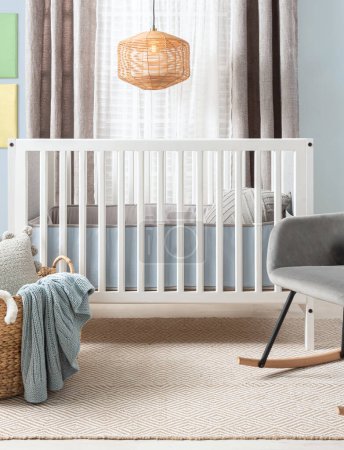 Heiter gestyltes Babyzimmer mit einer weißen modernen Krippe, einer plüschigen grauen Wippe und einem beigefarbenen Teppich mit Mustern. Das weiche Tageslicht filtert durch hauchdünne Vorhänge neben einem gewebten Rattan-Pendellicht.