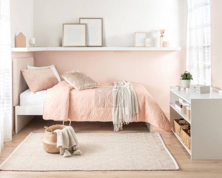 Bezauberndes Mädchenschlafzimmer, rosafarbenes Einzelbett mit gesteppter Bettdecke, bequemer Elfenbeinstrickwurf, flauschiges Kissen, geflochtener Quastenkorb, auf einem Musterteppich, unter einem weißen minimalistischen Regal, schickes Dekor.