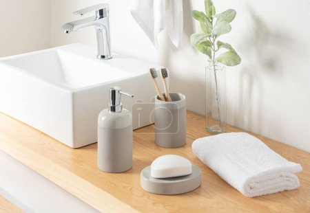 Detalle contemporáneo del baño eco-consciente, un elegante lavabo blanco cuadrado, cepillos de dientes de bambú natural en un soporte gris, un dispensador de jabón, con una toalla blanca de felpa en una encimera de madera caliente.