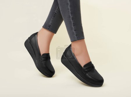 Elegant black comfort loafers worn by a woman in a standing pose, featuring sleek leather with a subtle decorative strap, complemented by stylish gray cropped leggings, against a soft beige background