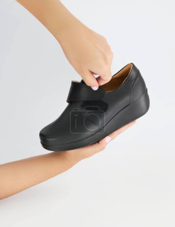Elegant black orthopedic shoe securely held in the hands of a young woman, a practical Velcro strap for easy adjustment and a supportive, comfortable sole design, for daily wear, white background.