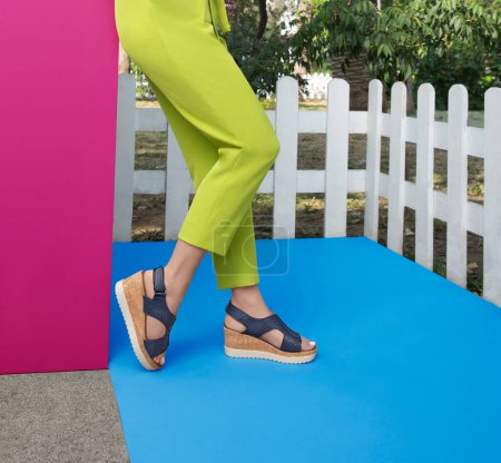 Outdoor scene featuring a woman modeling stylish navy blue wedge sandals, cork soles and perforated straps. stands on a bright blue floor, pink wall, wearing lime cropped pants, greenery background.