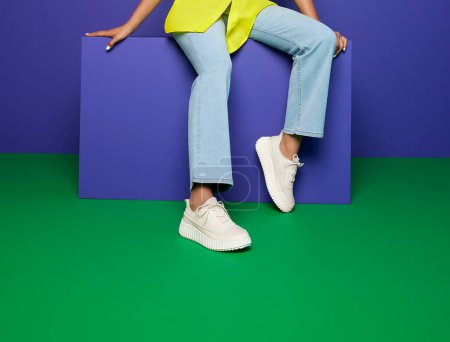 Young woman modeling trendy cream-colored lace-up sneakers with a chunky tread sole, seated on a purple block, a green and purple background. dressed in light blue jeans, yellow top, casual footwear.