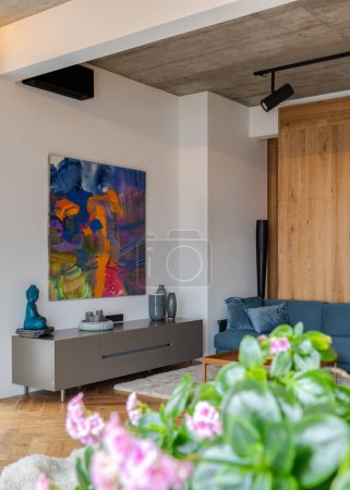 Foto de Un diseño interior con una sala de estar con un sofá azul, una pintura en la pared y una planta para mayor estética. La habitación está muy bien iluminada con iluminación ambiental y tiene suelos de madera - Imagen libre de derechos
