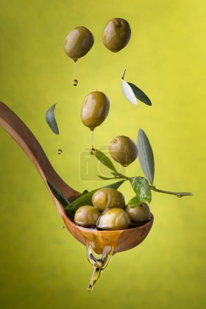 Foto de Cucharón con aceitunas y aceite sobre un fondo verde. Aceitunas, aceite de oliva virgen extra y hojas de oliva flotan en el aire - Imagen libre de derechos