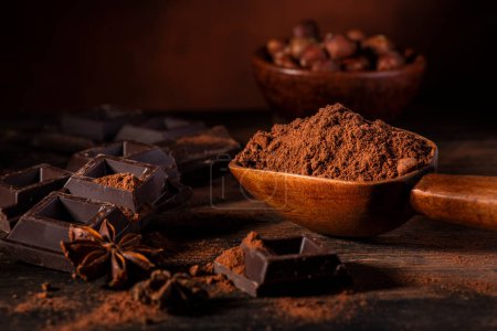Photo for In the foreground, in a dark environment, cocoa powder, pieces of dark chocolate and hazelnuts - Royalty Free Image