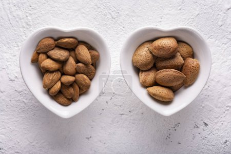 Photo for Top view, extreme close-up, against a textured white concrete background, two heart-shaped bowls with almonds in the shell and without. - Royalty Free Image