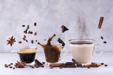 Foto de Composición animada con espresso, café con leche y capuchino en tazas de vidrio transparente entre especias aromáticas flotantes, trozos de chocolate y granos de café - Imagen libre de derechos