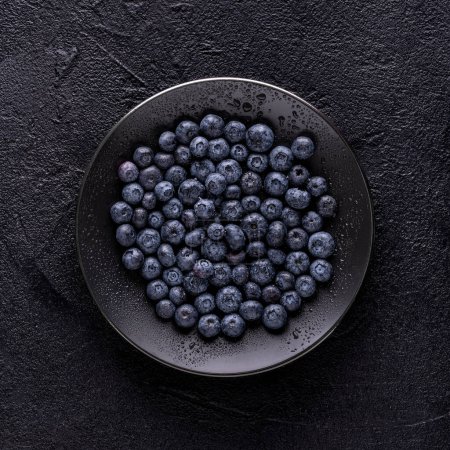 Photo for From above, on a black textured background, a black plate with fresh wet blueberries. - Royalty Free Image