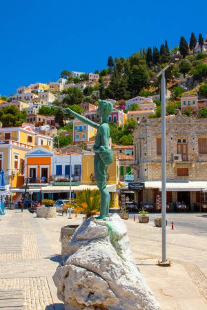 Foto de SYMI, Grecia - JUN 03, 2021. Escultura de niño en el paseo marítimo de Symi en el puerto frente a coloridas villas que se elevan por las empinadas laderas de las montañas. - Imagen libre de derechos