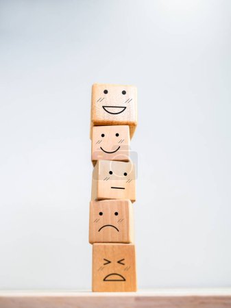 Évaluation du service à la clientèle, rétroaction et sondage sur la satisfaction des employés. Émoticône souriant heureux sur un autre visage émotionnel, empilant des blocs de bois sur fond blanc, style vertical.
