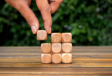 Last piece wood cube block in hand putting on wooden cube as puzzles stacking arranging completed on table and green plant background. Solution, solve problem, business goal and success plan concepts.