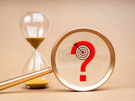 Problem analysis and solving, time to making right decision concept. Red question mark icon with target symbol in big magnifying glass lens near golden hourglass on light brown background.