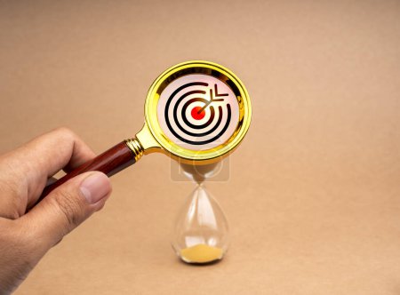 Photo for Short-term business goals encompass work that helps an organization reach its mid-term goals concept. Target icon in gold magnifying glass lens in hand while zoom in at hourglass on brown background. - Royalty Free Image