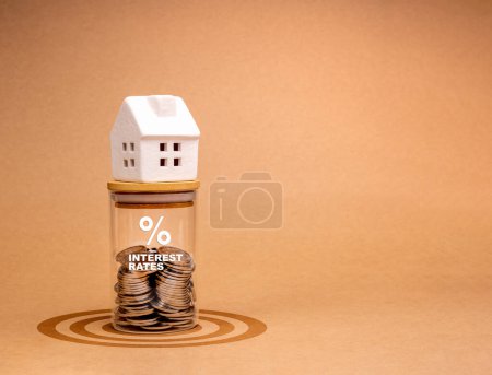 Hypothekenzinsen, Eigenheimsteuer, Immobilienfinanzierungskonzepte. Weißes Miniaturhaus auf Glasflasche mit Prozentsymbol und Text "INTERESSE RATES", Münzen innen, auf Zielsymbol, brauner Hintergrund.