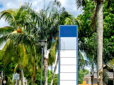 Banner vacío blanco y azul en blanco o letrero de la calle para la publicidad junto a la carretera cerca de las palmeras y el jardín verde. Mockup alto y alto letrero al aire libre para el logotipo de la tienda o tienda en el parque público de la ciudad.