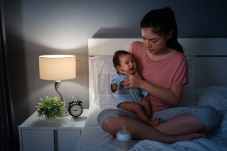 Foto de Madre sosteniendo el eructo y llorando bebé después de alimentar la leche en una cama por la noche - Imagen libre de derechos