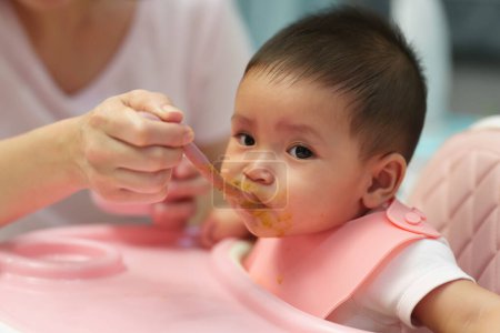 Photo for Mother feeding food to her infant baby eating with a spoon at home - Royalty Free Image