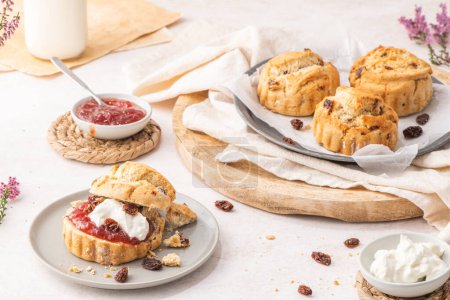 Photo for Traditional English pastries for afternoon tea: scones. Homemade raisin scones with clotted cream and homemade strawberry jam. Scones on plate served with milk on table - Royalty Free Image