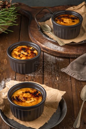 Creme brulee. Bowls with French vanilla cream dessert with caramelised sugar on top, spoons, dark rustic table. Leite creme, portuguese desert similar to creme brulee, cream brulee and burnt cream.
