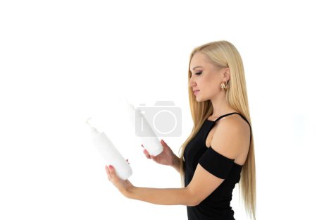 Téléchargez les photos : Une belle femme blonde en robe noire aux cheveux parfaitement raides sur un fond blanc isolé choisit un produit de soin pour les cheveux lissés. Vue latérale - en image libre de droit
