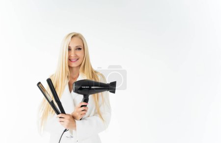Téléchargez les photos : Femme européenne élégante coiffeur souriant et tenant sèche-cheveux noir et fer à friser dans ses mains.Isolé sur fond blanc, concept de soins capillaires - en image libre de droit