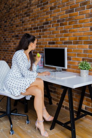 Photo for A stylish European girl is sitting on a white chair near a desktop with a PC. Place for text, concept of calls, orders, receiving orders, form of delivery - Royalty Free Image