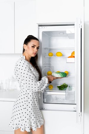 Téléchargez les photos : A European girl confectioner stands in a white kitchen in a white polka-dot dress near an open refrigerator in her hands holding a mousse cake - en image libre de droit