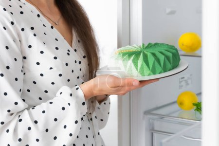 Téléchargez les photos : A girl holds a mousse cake with a live flower cake in her hands near an open refrigerator. The concept of food storage, confectioner, sweets - en image libre de droit