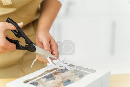 Téléchargez les photos : A European confectioner in a bright kitchen in an apron cuts the tape for packaging cupcakes with scissors - en image libre de droit