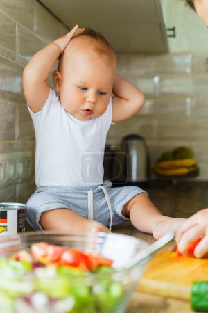 The baby is sitting on the kitchen table. Nearby, mom is cutting food into salad. The boy reacts emotionally. High quality photo