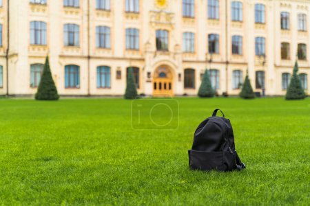 Téléchargez les photos : Un sac à dos d'école noir debout sur de l'herbe verte sur le fond d'une école, une université. Photo de haute qualité - en image libre de droit