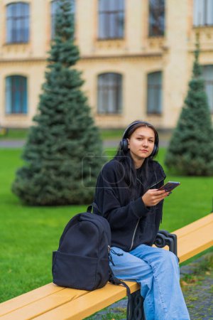 A female student in headphones sits on a bench near the university with a phone in her hands, the first of September, the academic year begins. High quality photo