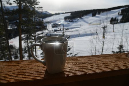 Téléchargez les photos : Cup of fresh morning coffee on the porch of the condo at ski resort, blurry snow slopes on the background. - en image libre de droit