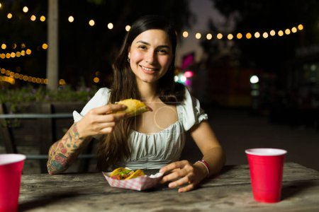 Foto de Hermosa mujer hispana pidiendo comida callejera mexicana desde el camión de comida y feliz de comer deliciosos tacos - Imagen libre de derechos