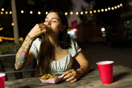 Attractive hispanic woman looking hungry enjoying eating delicious mexican tacos from the food truck at night