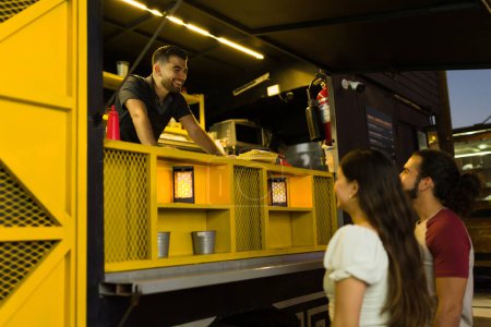 Foto de Emocionado trabajador de camiones de comida riendo y mirando feliz hablando con los clientes que ordenan comida rápida - Imagen libre de derechos