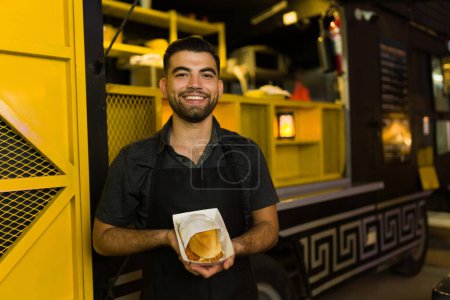 Foto de Chef latino alegre en el camión de comida listo para servir comida rápida y mostrando un delicioso sándwich - Imagen libre de derechos