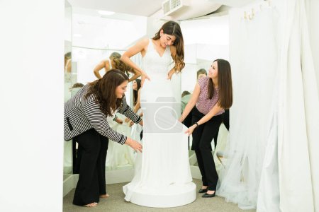 Téléchargez les photos : Magnifique jeune femme essayant une robe de mariée dans la loge avec l'aide de ses amies - en image libre de droit