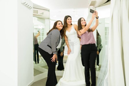 Téléchargez les photos : Joyeuses amies et demoiselles d'honneur prenant un selfie avec la future mariée essayant sa robe de mariée au magasin de mariée - en image libre de droit