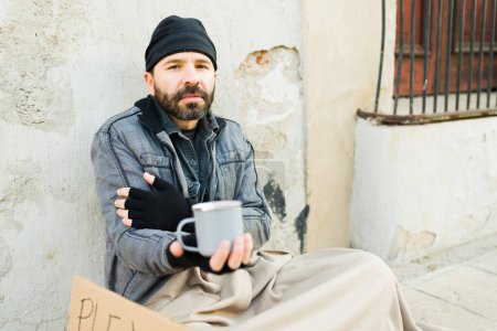 Hombre hambriento triste sin hogar haciendo contacto visual mientras sostiene una taza pidiendo dinero y sintiendo frío