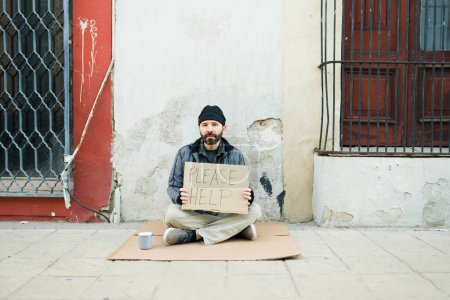 Photo for Portrait of a homeless man in poverty with a carboard sign asking for help while living on the streets and looking at the camera - Royalty Free Image