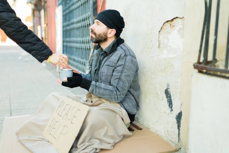 Foto de Mendigo sin hogar sintiendo hambre recibiendo dinero en su taza de un hombre mientras vive en las calles - Imagen libre de derechos
