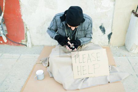 Photo for Poor homeless man counting the coins money to buy food while sitting on a carboard with a sign of please help - Royalty Free Image
