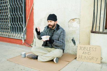 Foto de Hombre enfermo sin hogar comiendo una sopa y hablando solo mientras vive en las calles - Imagen libre de derechos