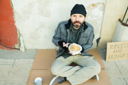 Foto de Pobre hombre hambriento sin hogar haciendo contacto visual mientras come una sopa caliente en la calle - Imagen libre de derechos