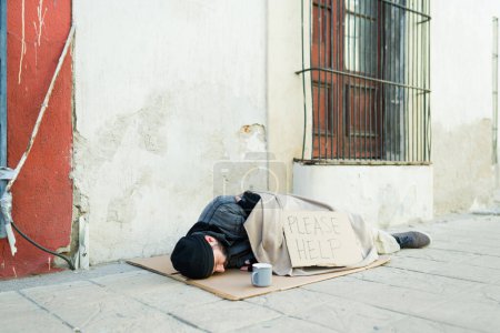 Foto de Hombre sin hogar mendigo frío cubierto con una manta que duerme en la calle con un signo de cartón por favor ayuda - Imagen libre de derechos