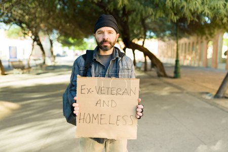 Téléchargez les photos : Ex-vétéran et sans-abri vivant dans la pauvreté dans la rue tenant une pancarte en carton demandant de l'aide tout en luttant - en image libre de droit