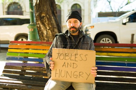 Foto de Triste mendigo sin hogar sentado en el banco del parque y sosteniendo un cartel de cartón sin trabajo y hambriento - Imagen libre de derechos